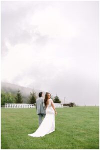 Bride and groom walking away from the camera on a green lawn with misty mountains in the background at Overlook Barn in Banner Elk, NC.