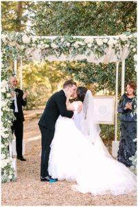 Bride and groom share their first kiss under a floral arch at Vanlandingham Estate, Charlotte, NC.